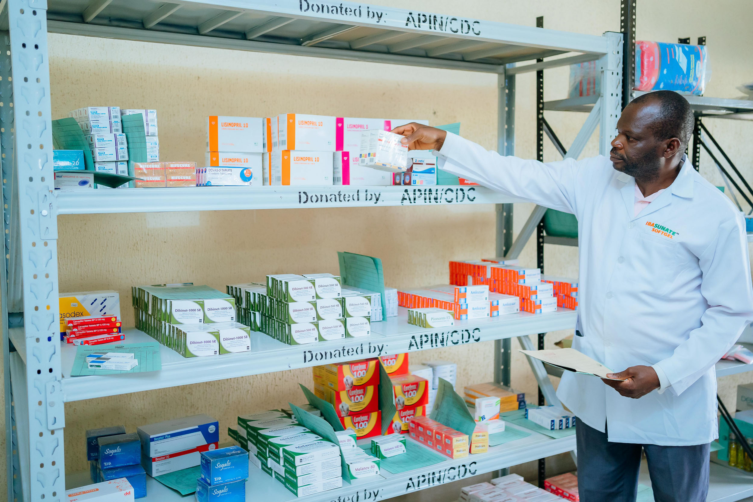 Pharmacist inspecting pharmaceutical stocks and health commodities on shelving marked Donated by APIN/CDC. 
